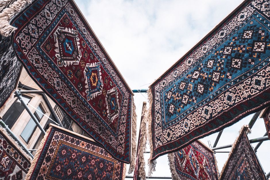 Inside a bustling market at Brixton Village, textile items including colorful woven blankets and carpets are displayed on tables and hanging on a clothesline under a high, industrial-style roof with m