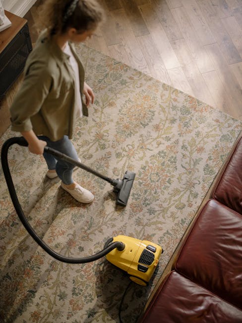 A yellow vacuum cleaner with a black hose attachment resting on a patterned area rug in a living room. The room features a wooden floor and a black metal and wood media console against the wall, which