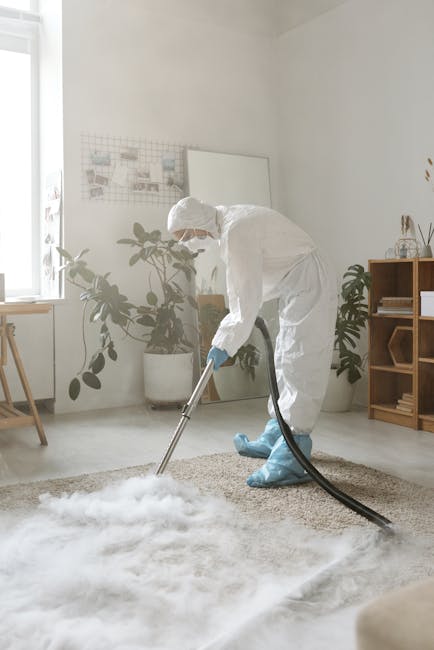 A professional cleaner wearing a white protective suit, face mask, and blue gloves vacuuming a cream-colored carpet in a bright, modern living room. The room features white walls, a wooden shelf with 