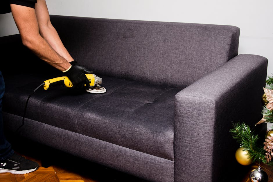 A person using a handheld electric polisher for surface cleaning on a dark grey fabric sofa with a cushioned backrest and armrest, situated in a well-lit living room. The sofa is positioned on a woode